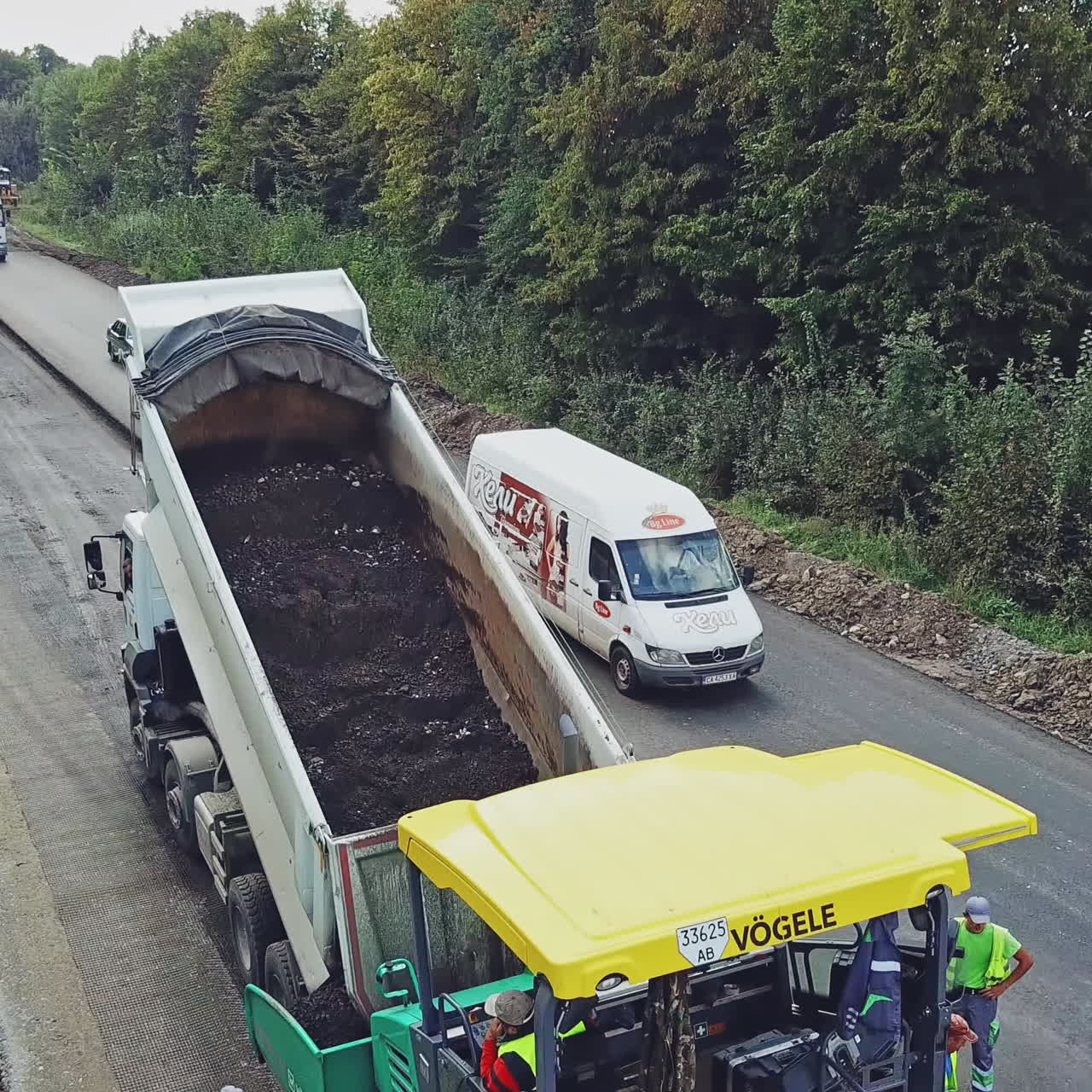 A truck tosses over bitumen into a wide paver to lay a new roadway surrounded of repairmen. Road construction equipment. Aerial view.