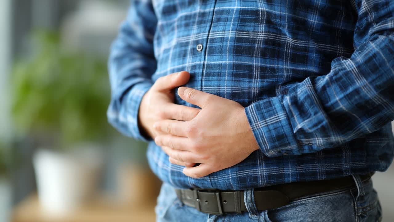 Un hombre con camisa de cuadros azul experimentando malestar, sujetándose el estómago con ambas manos, indicativo de posible indigestión o problemas gastrointestinales en un ambiente informal.