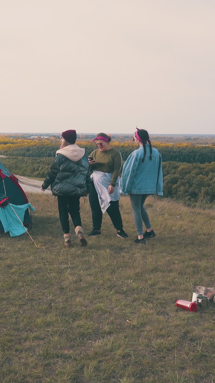 young girls tourists listen to music dancing at blue tent on green hill top against pictorial calm river at sunset