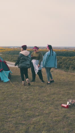 young girls tourists listen to music dancing at blue tent on green hill top against pictorial calm river at sunset