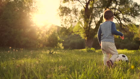 Boy-standing-with-a-soccer-ball-in-the-summer-running-on-the-field-with-grass-rear-view