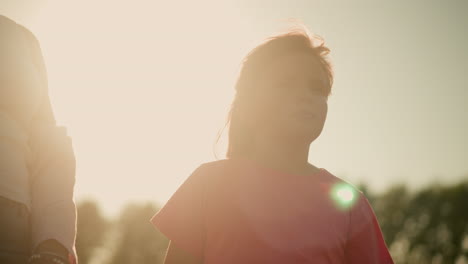 little girl in pink shirt looking thoughtful under bright sunlight with partial view of adult standing near her, sunlight creates glowing effect around them, giving scene warm and serene vibe