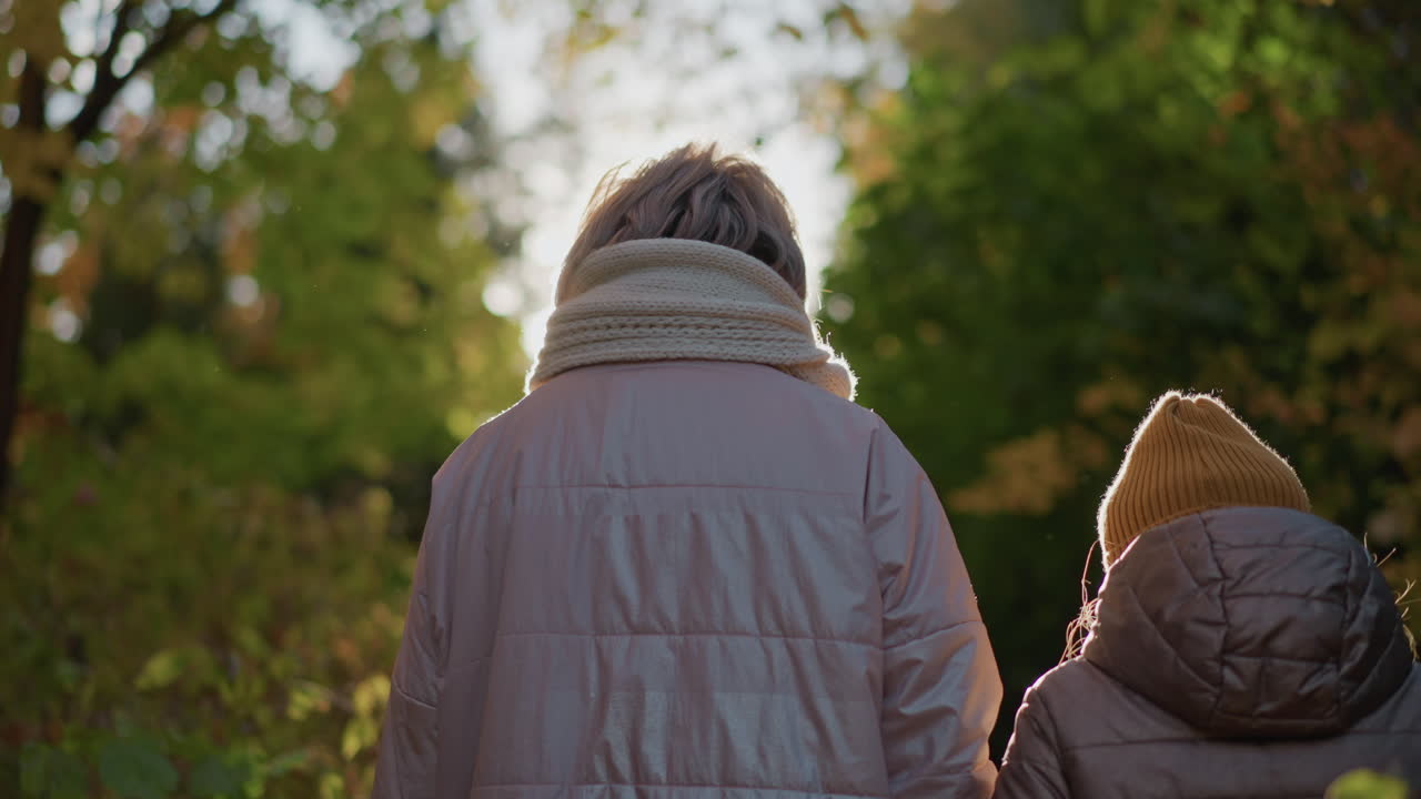 back view of mother and child walking along forest trail bathed in golden sunlight glow, autumn foliage framing path, winter jackets and scarves signaling chilly air, warm family