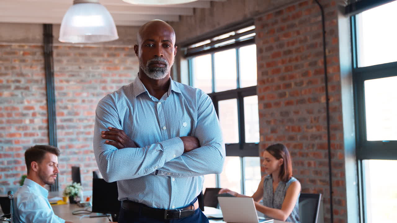 Portrait Of Serious Mature Businessman Standing In Busy Office With Colleagues Working In Background