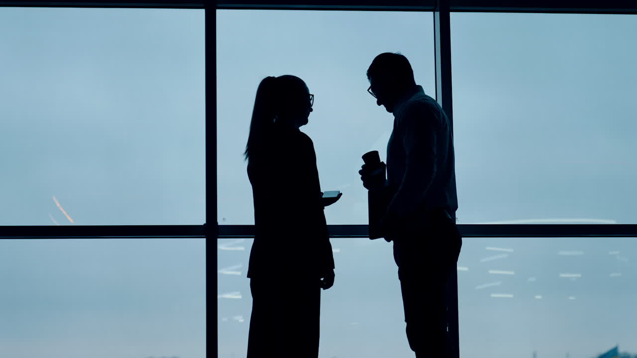 Cropped silhouettes of two people standing at the window. Man and woman standing in the office at window look at smartphone.