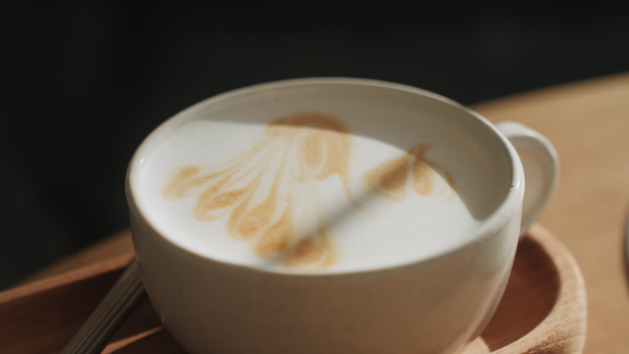 Xícara de latte em bandeja de madeira aconchegante, banhada pela luz solar quente, com delicadas pétalas de arte em espuma flutuando na crema cremosa, caneca de cerâmica em pires com colher, sombra suave cruzando a superfície, criando um ambiente de café íntimo e tátil.