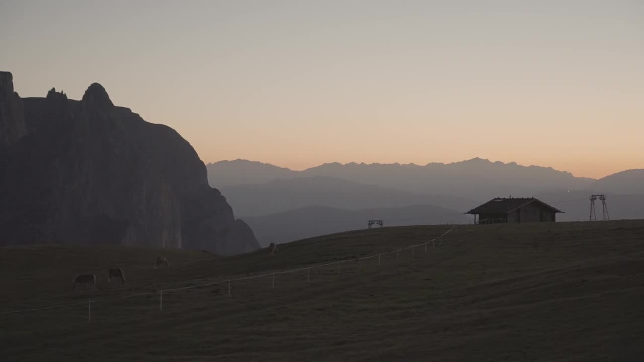 Serene pasture in South Tyrol with wooden cabin and grazing cows at sunrise, surrounded by rolling hills and distant mountain peaks under a warm morning sky