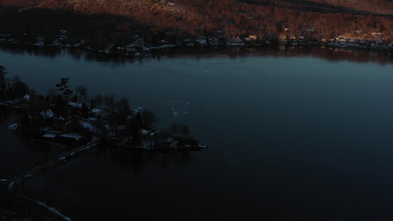 Dlouhý přelet dronu nad zimním jezerem a jezerními domy směrem k kopcům a lesu při východu slunce.