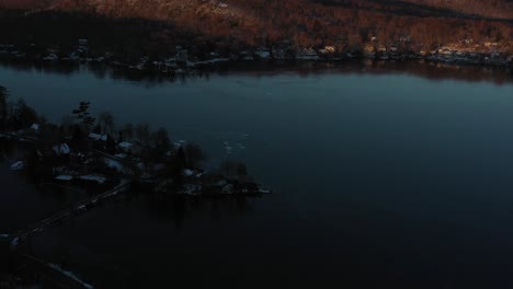 Long-drone-flyover-winter-lake-and-lake-houses-towards-hills-and-forest-at-sunrise