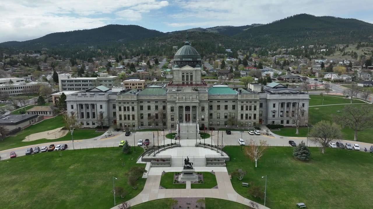 La cupola del Campidoglio dello Stato del Montana si erge sopra lo skyline di Helena, incorniciata da edifici governativi, strade alberate e montagne boscose sotto un cielo nuvoloso. Helena, Montana, USA. Ripresa aerea con drone in allontanamento.