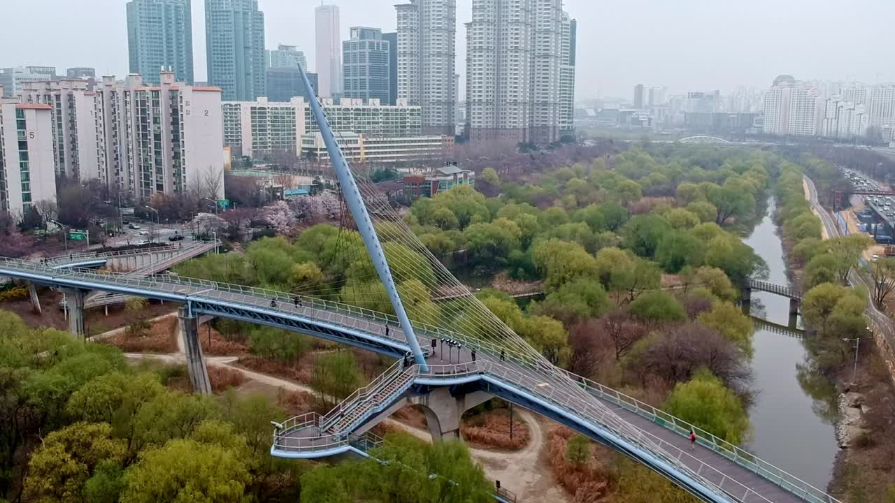 Rightward glide shot of an artfully designed bridge over a green space, centered by a skyline and a busy road.