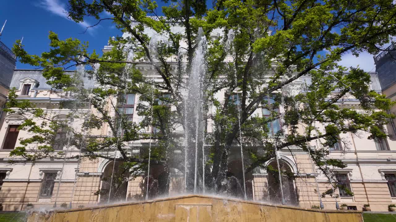 Vista frontal en cámara lenta de la fuente del Ayuntamiento de Iasi con chorros de agua y ramas de árboles enmarcando un edificio histórico en un día soleado
