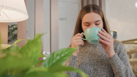 Young lady enjoys hot chocolate drink, gently lifting cup to lips then breathing out satisfied, cozy indoor space with flower decor and lamp stand in soft background adds warmth and relaxation