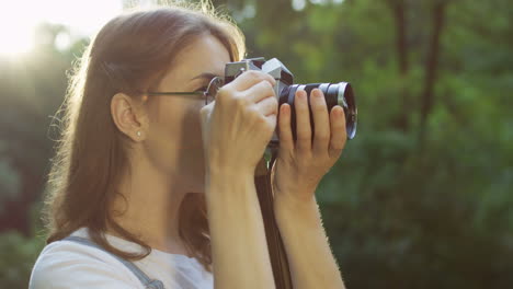 古いビンテージ写真カメラで公園で写真を撮るメガネの美しい若い女性の側面図