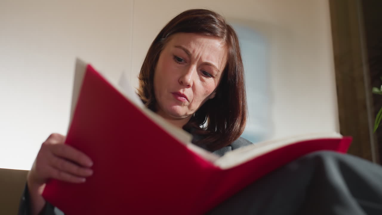 Closeup of businesswoman with serious expression reading red folder while seated in modern office, deeply focused on reviewing important paperwork or legal documents under natural daylight