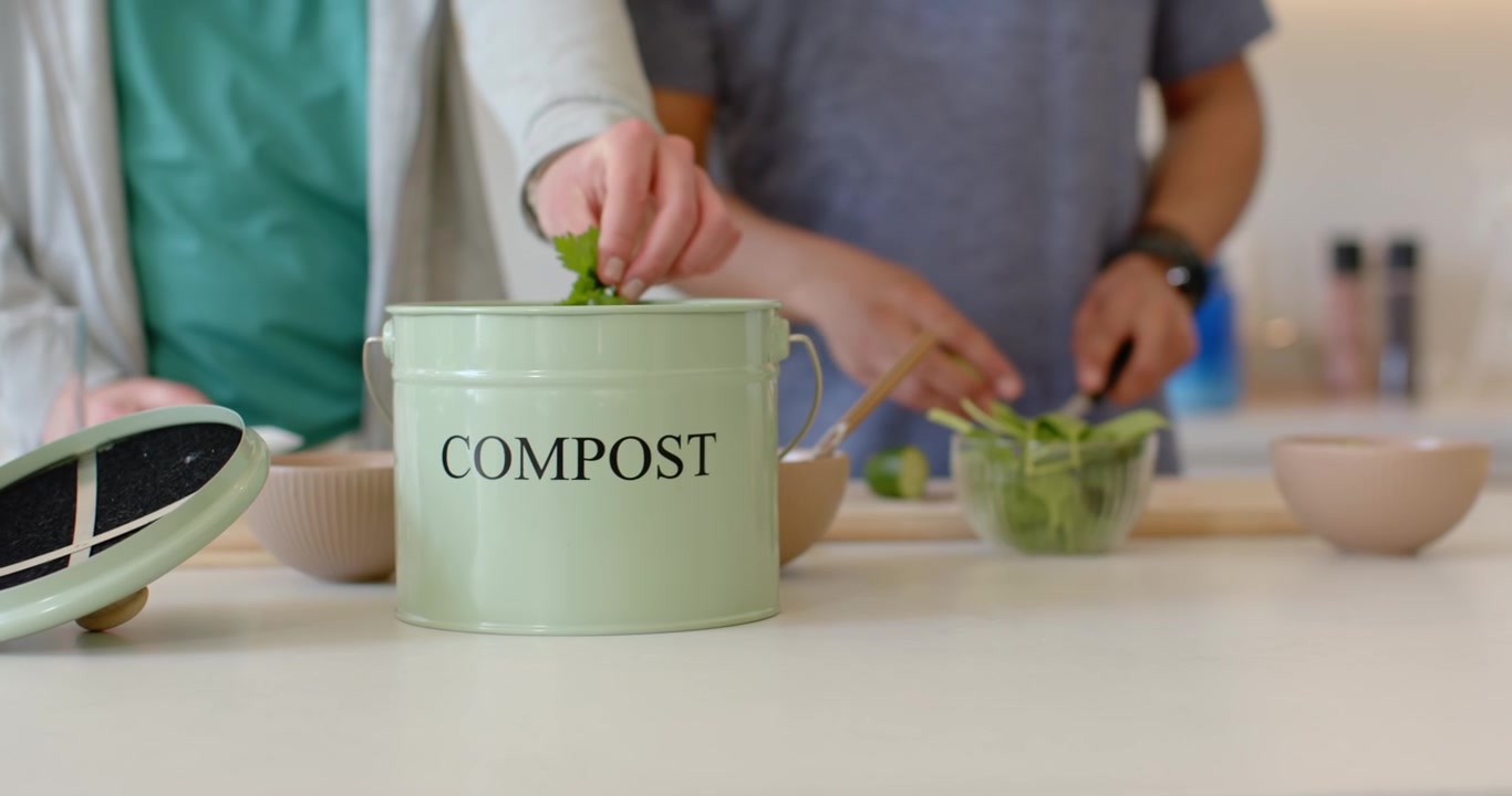 Un couteau coupe des légumes pour salade, des mains jettent des restes dans un seau à compost vert pâle sur le comptoir