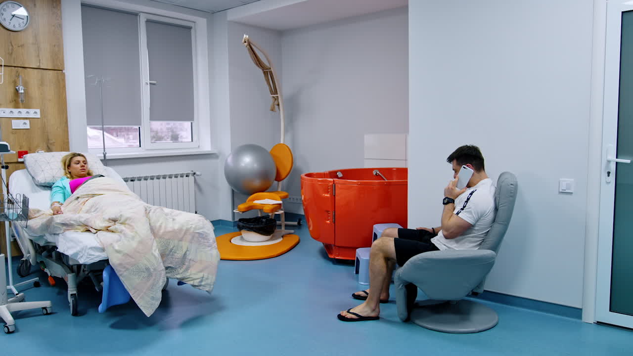 Pregnant Caucasian woman lies in the bed at maternity hospital. Husband sits in an armchair speaking on the phone.