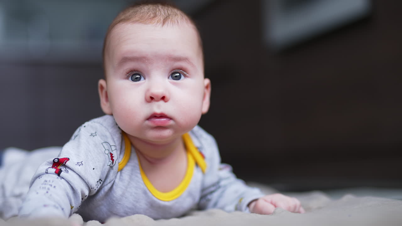 Beautiful Caucasian child lies on his tummy holding head up high. Curious little boy looks sideways with eyes wide open. Close up.
