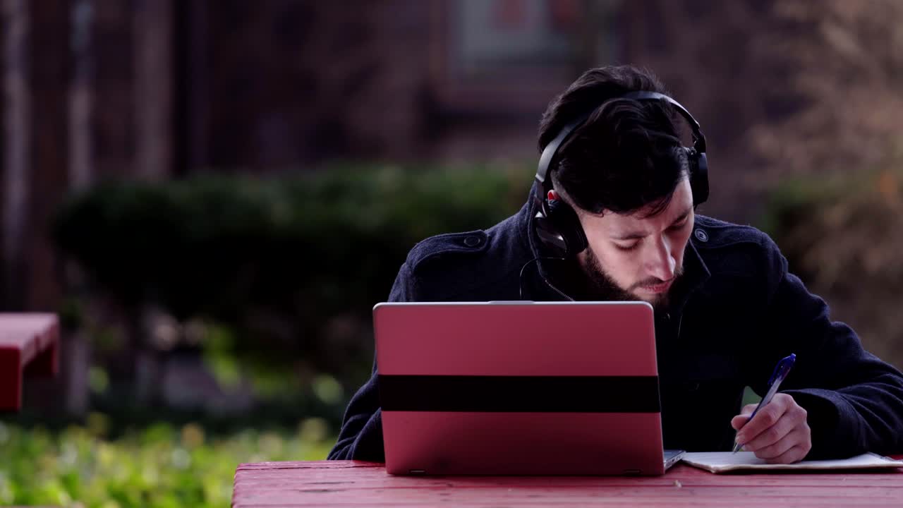 Mann med svart jakke skriver notater på laptopen mens han hører på musikk - Nærbilde