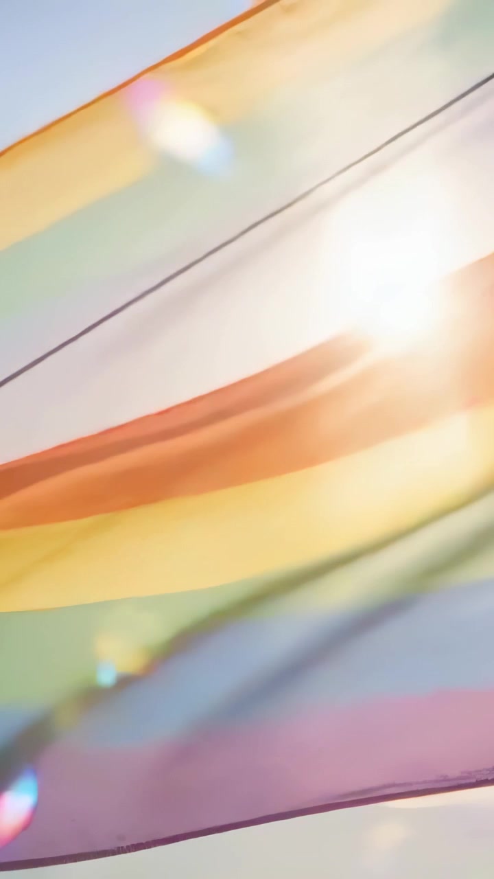 Vertikales Video: Eine Regenbogenflagge weht im Wind an einem schrägen Seil vor dem Himmel, die Sonne scheint mit Lichtreflexen.