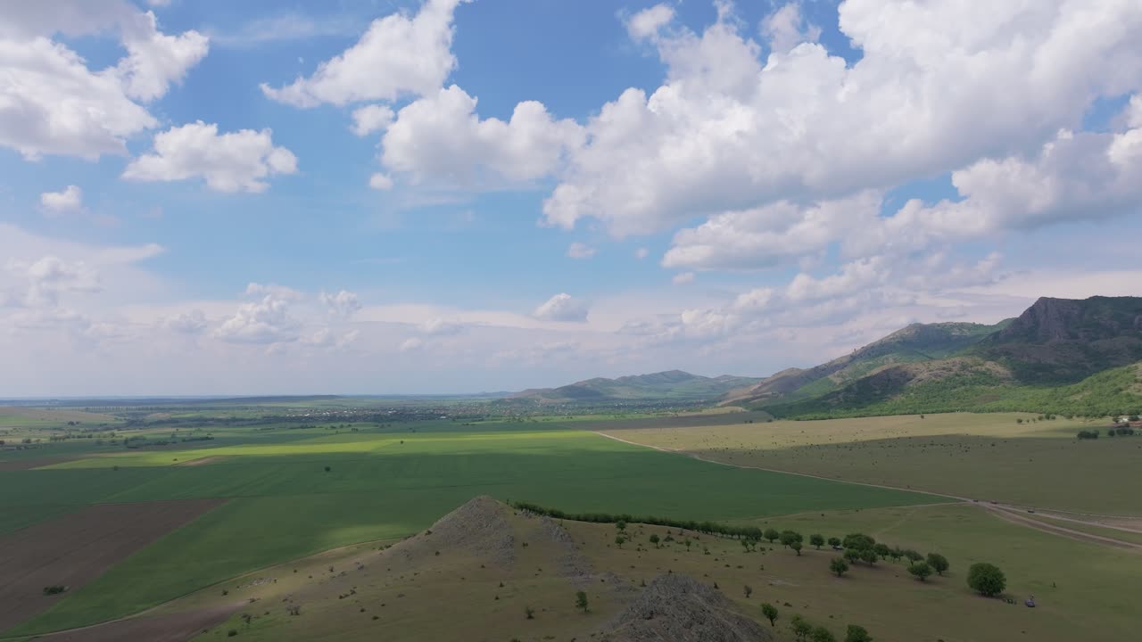 Vast green agricultural fields under a vibrant blue sky dotted with fluffy white clouds, framed by rugged mountains and sloping hills