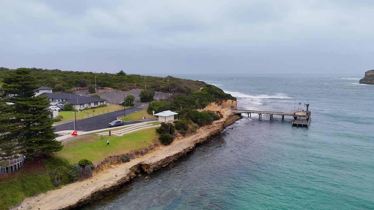 Aerial view of Port Campbell's pier and coastline, showcasing lush greenery, ocean waves, and overcast skies captured by a drone