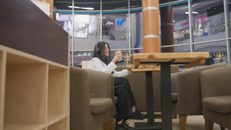 side view of young woman wearing headphones focusing on work at cafe table as she places paper coffee cup down with background featuring large glass panel and modern shopping mall reflections