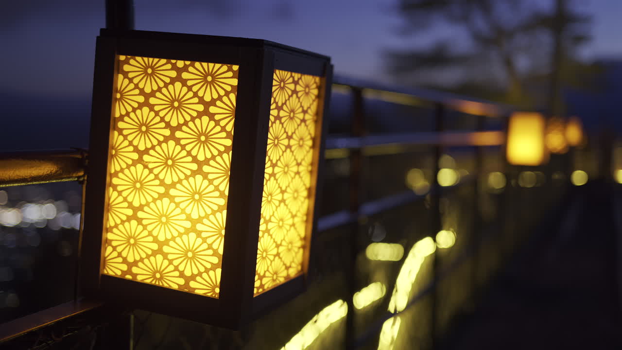 Warm orange Japanese lanterns cast a gentle glow along a walkway at dusk, creating a serene atmosphere as the city skyline sparkles in the background, inviting evening strolls