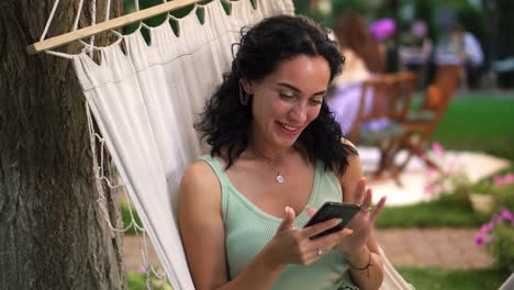 a girl with curly hair sits in a hammock outdoors, shows her smartphone, clicks on the screen