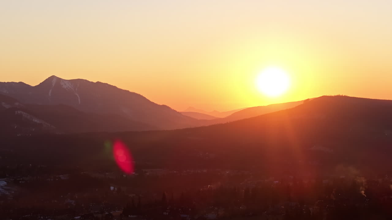 Sun dips below mountain ridge casting warm orange glow over the silhouetted terrain, aerial tracking left