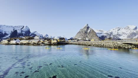 Kayakistas-En-Aguas-Serenas-En-Lofoten,-Noruega,-Con-Montañas-Nevadas-Bajo-Un-Cielo-Azul-Claro,-Plano-Amplio