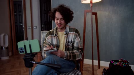 a happy brunette guy with curly hair watches videos and blogs using a smartphone and makes notes in his notebook while sitting on the sofa at home in a modern apartment