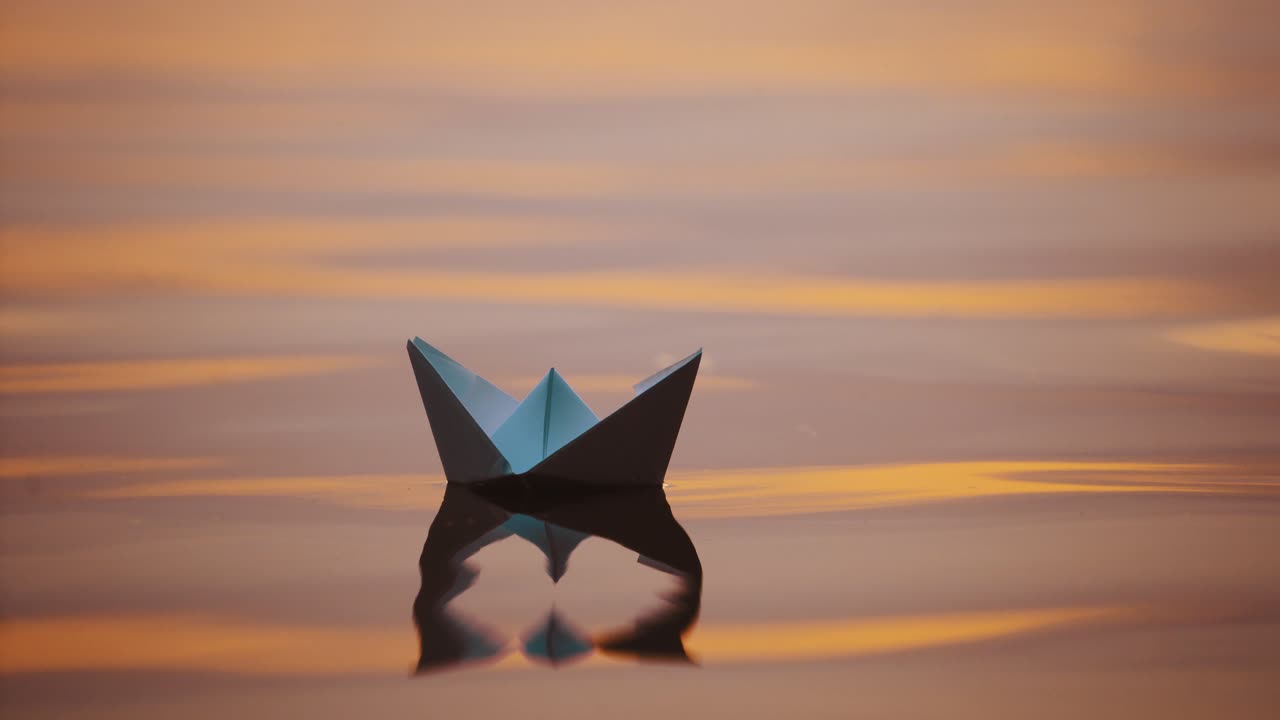 Origami boat on water surface in the evening. White paper boat swims with its shadow reflection in water. Close-up.