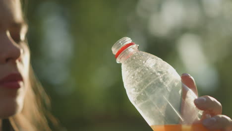 close-up of hand holding clear plastic bottle with condensation and some liquid, the bottle tilted towards sunlight, creating soft glow in background, with blurred greenery