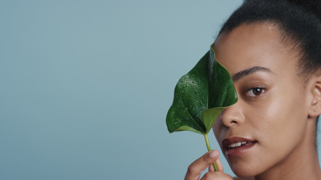 retrato de una hermosa mujer afroamericana tocando la cara con hojas acariciando una piel suave y saludable disfrutando de una belleza natural juguetona sobre un fondo azul concepto de cuidado de la piel