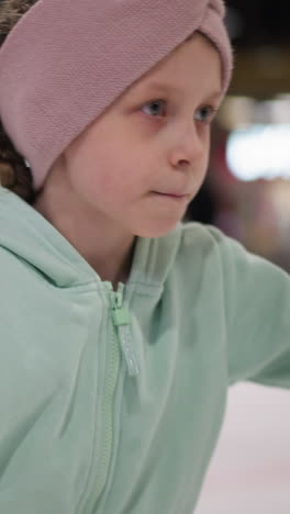 primer plano de una dama con una sudadera verde menta y una cinta rosa para la cabeza, dirigiéndose atentamente a la cámara mientras patina sobre hielo, el fondo presenta luces borrosas y personas patinando