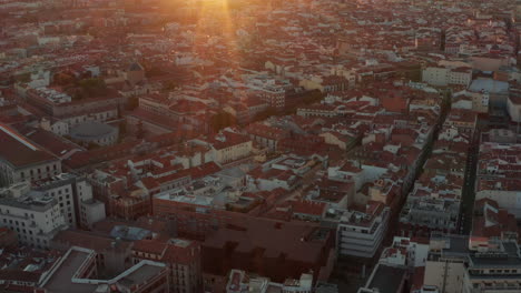 Fly-above-centre-of-large-city.-Aerial-panoramic-view-of-various-buildings-with-red-roofs.-Tilt-up-reveal-cityscape-and-setting-sun-on-horizon.