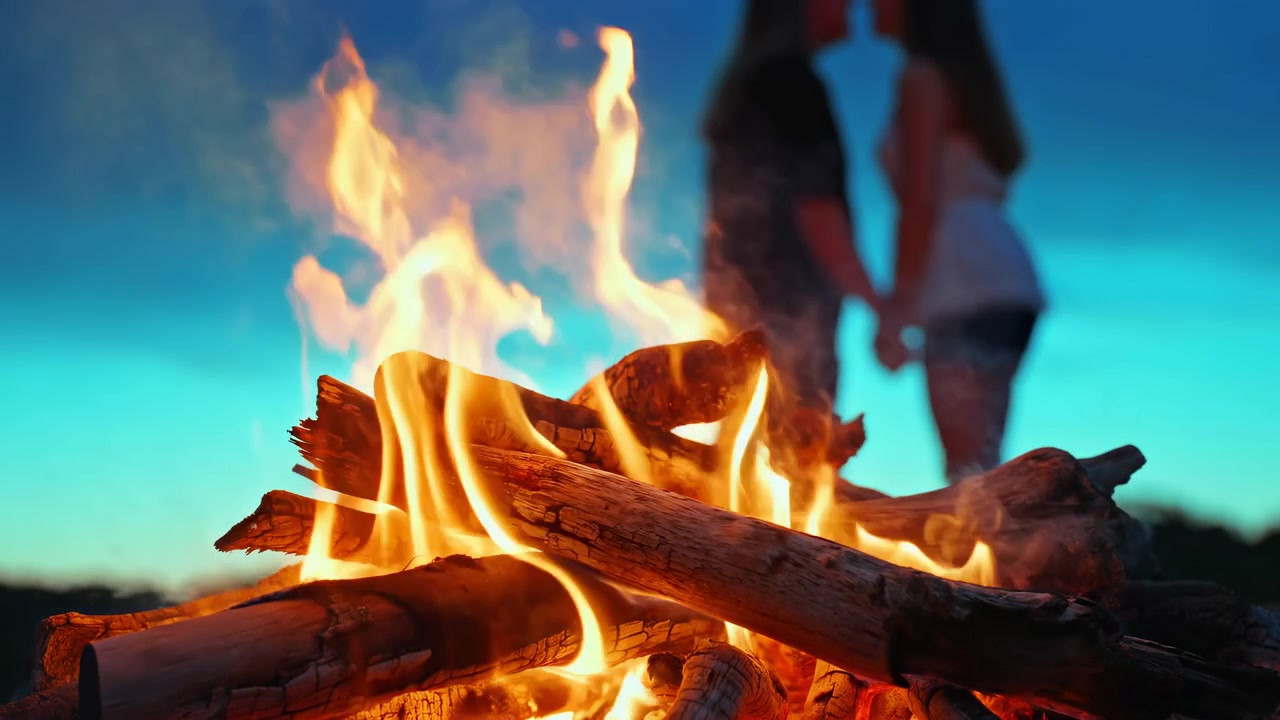 Niedrige Nahansicht von brennenden Holzscheiten mit hohen, sich kräuselnden Flammen, glühender Glut und aufsteigendem Rauch im scharfen Vordergrund, während verschwommene menschliche Silhouetten Hände vor einem türkisfarbenen Dämmerungshimmel und weichem Außenlicht halten.
