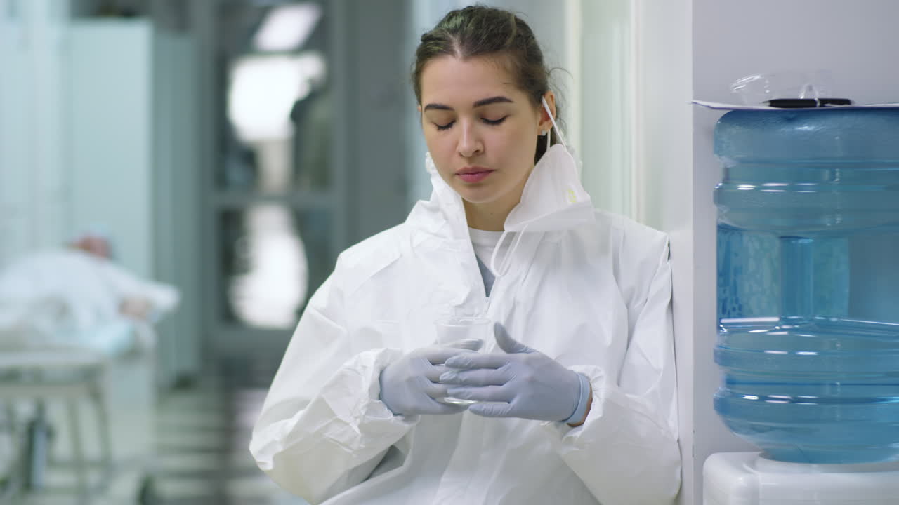 Joven médica con uniforme protector bebiendo agua