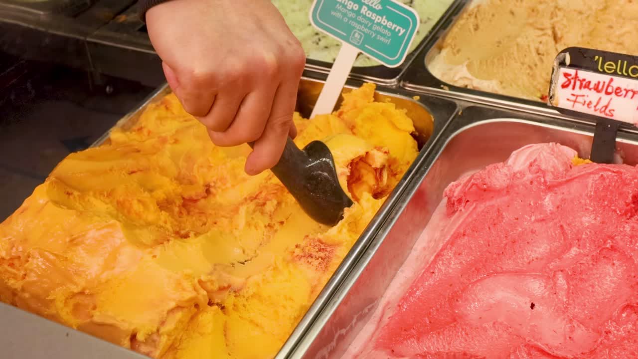 Close-up of a hand scooping colorful ice cream from a display with various flavors.