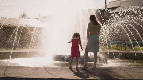 Una-Niña-Toca-El-Agua-De-Una-Fuente-Y-Toma-La-Mano-De-Su-Madre-En-El-Parque.