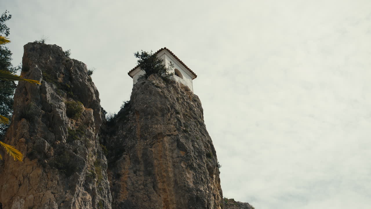 Small chapel tower perched on top of a steep rocky cliff in a dramatic mountain landscape. Minimalistic composition with rugged textures and a sense of height. In Guadalest, Spain,