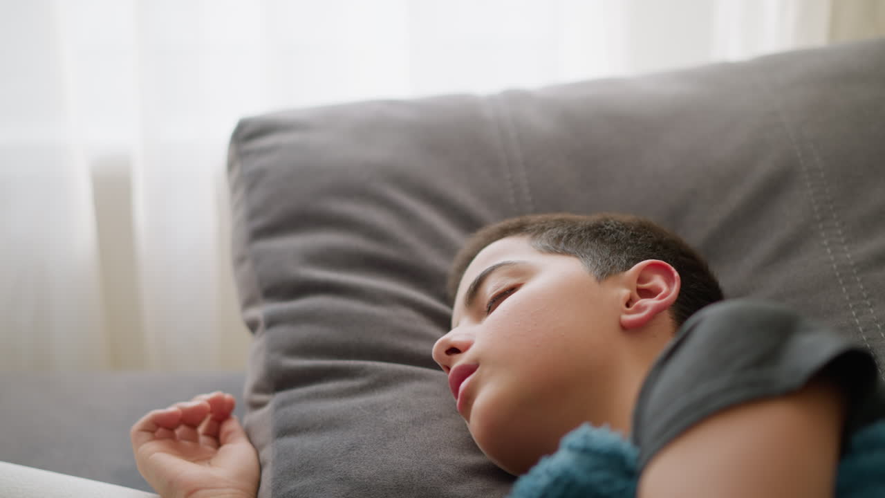 Close-up of young child resting on bed, covered with blue blanket, turning and placing hand on head in discomfort, showing signs of illness