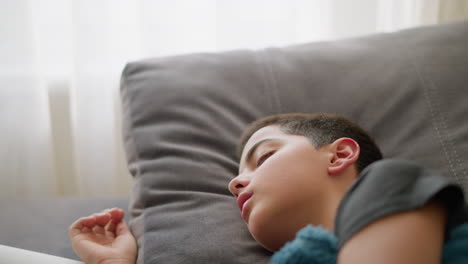 close-up of young child resting on bed, covered with blue blanket, turning and placing hand on head in discomfort, showing signs of illness