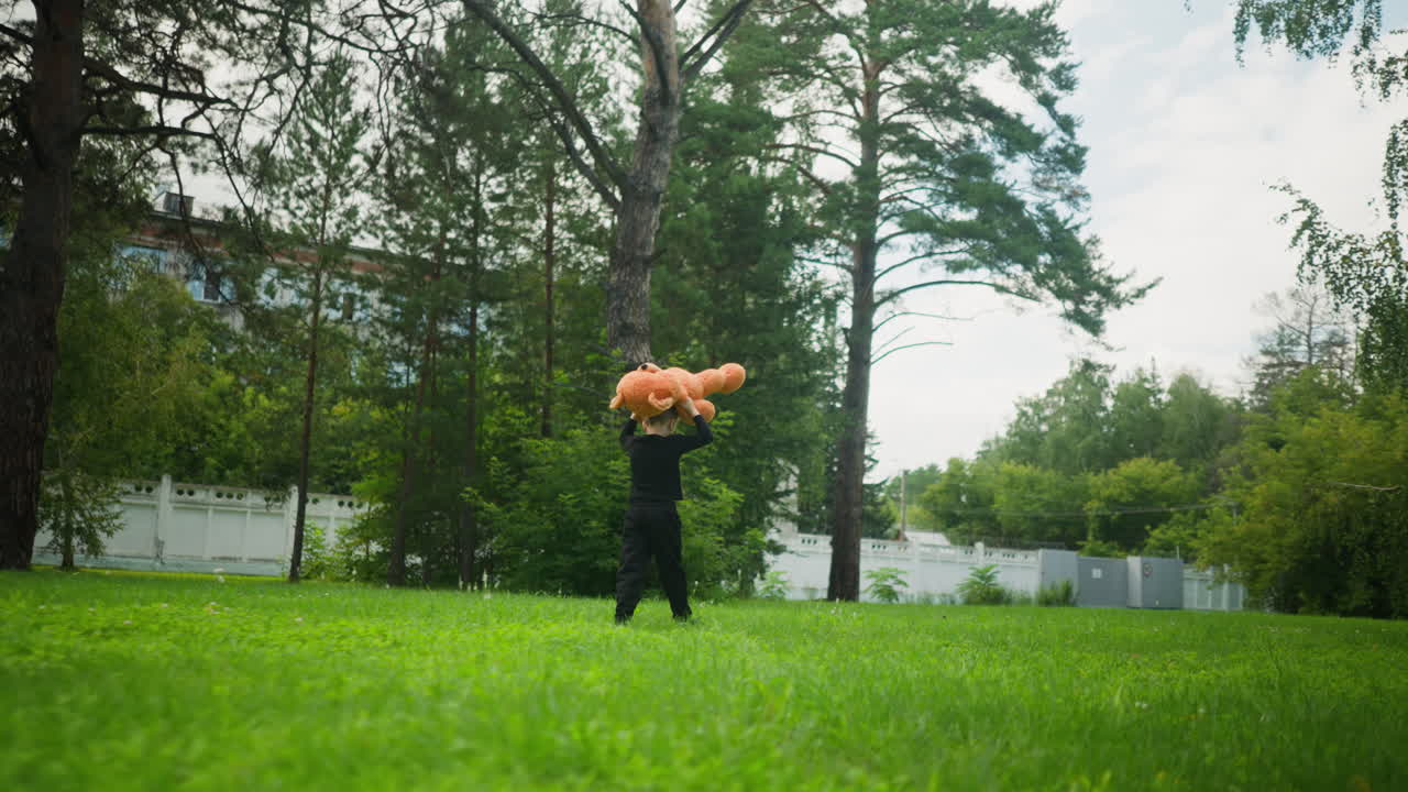 Young boy dressed in black walks across grassy field carrying teddy bear on head, surrounded by tall trees and distant buildings under overcast sky