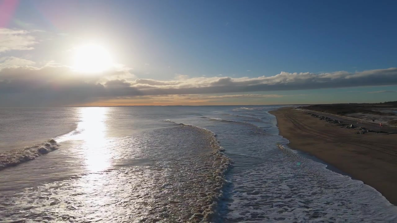 Luftfoto fanger strandscene i den gyldne time. Sollys reflekteres fra blide bølger, der møder den sandede kyst. Bløde klitter indrammer den rolige havhorisont. Den fredfyldte atmosfære fremkalder ro og afslapning.