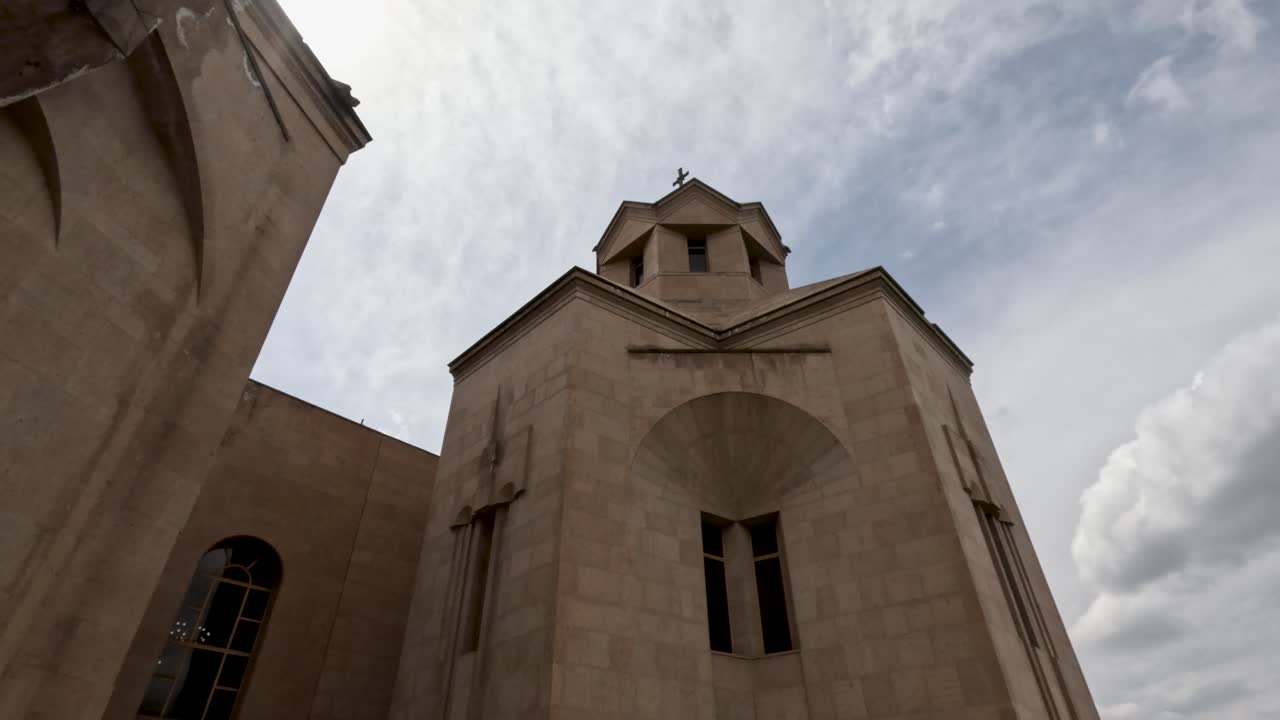 Un edificio ecclesiastico si erge imponente con le sue mura in pietra e una croce in cima. La telecamera cattura i dettagli della sua struttura e delle nuvole circostanti nella luce del pomeriggio.