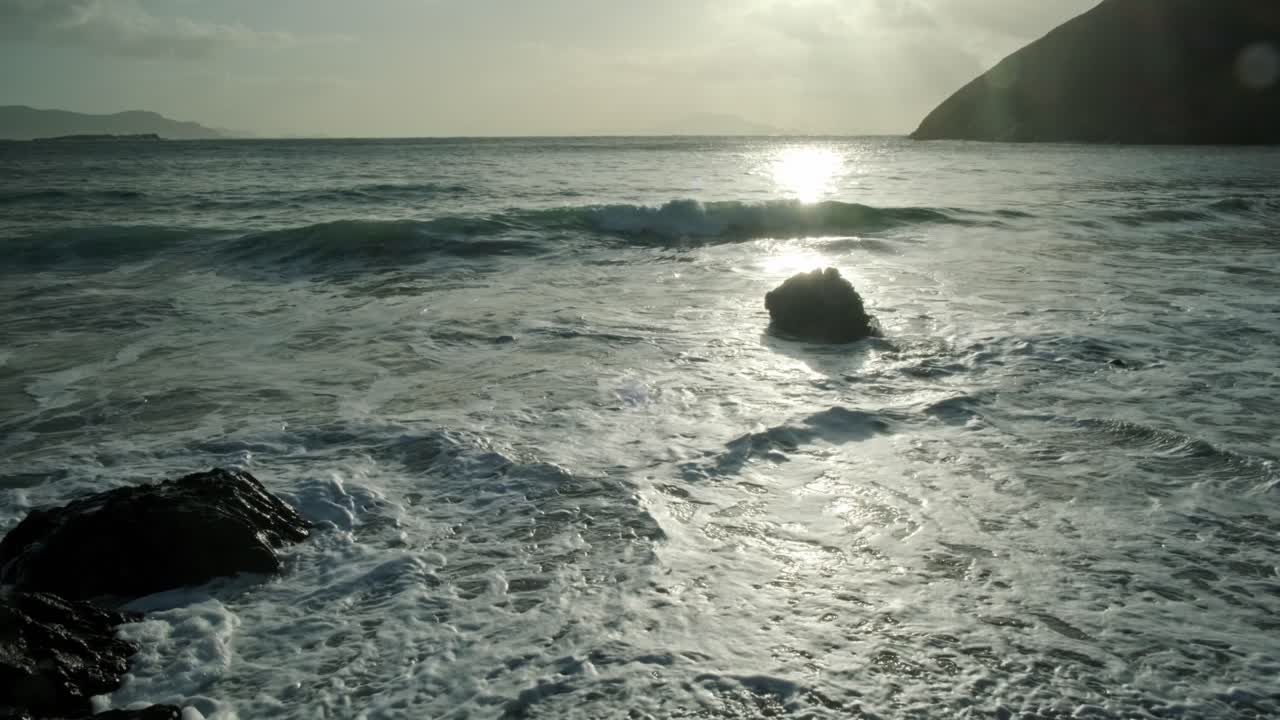 Wide shot of the ocean with beautiful backlight from the sun. Waves hitting on stone at the beach. Huge hill in the background.