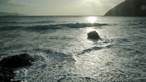 wide shot of the ocean with beautiful backlight from the sun. waves hitting on stone at the beach. huge hill in the background.
