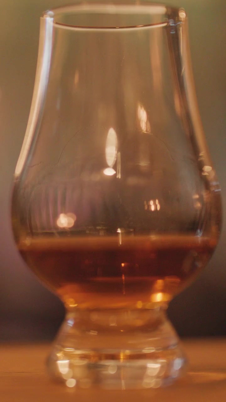 Un primo piano di whisky che vortica in un bicchiere a forma di tulipano su una superficie di legno, illuminato da una luce di candela soffusa e calda con uno sfondo sfocato.